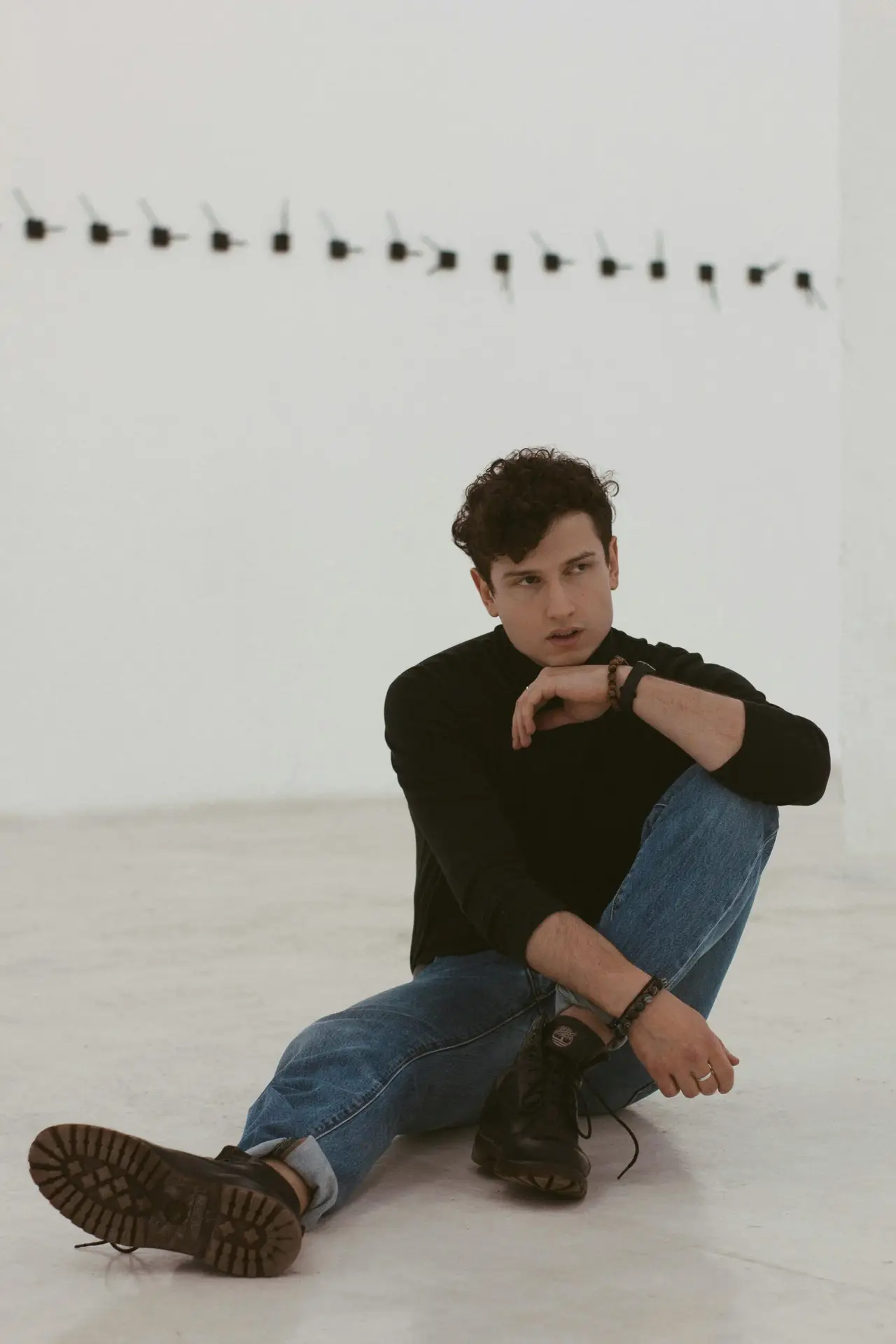 Hombre sentado posando con suéter negro, jeans azul arremangado, botas de cuero, y accesorios